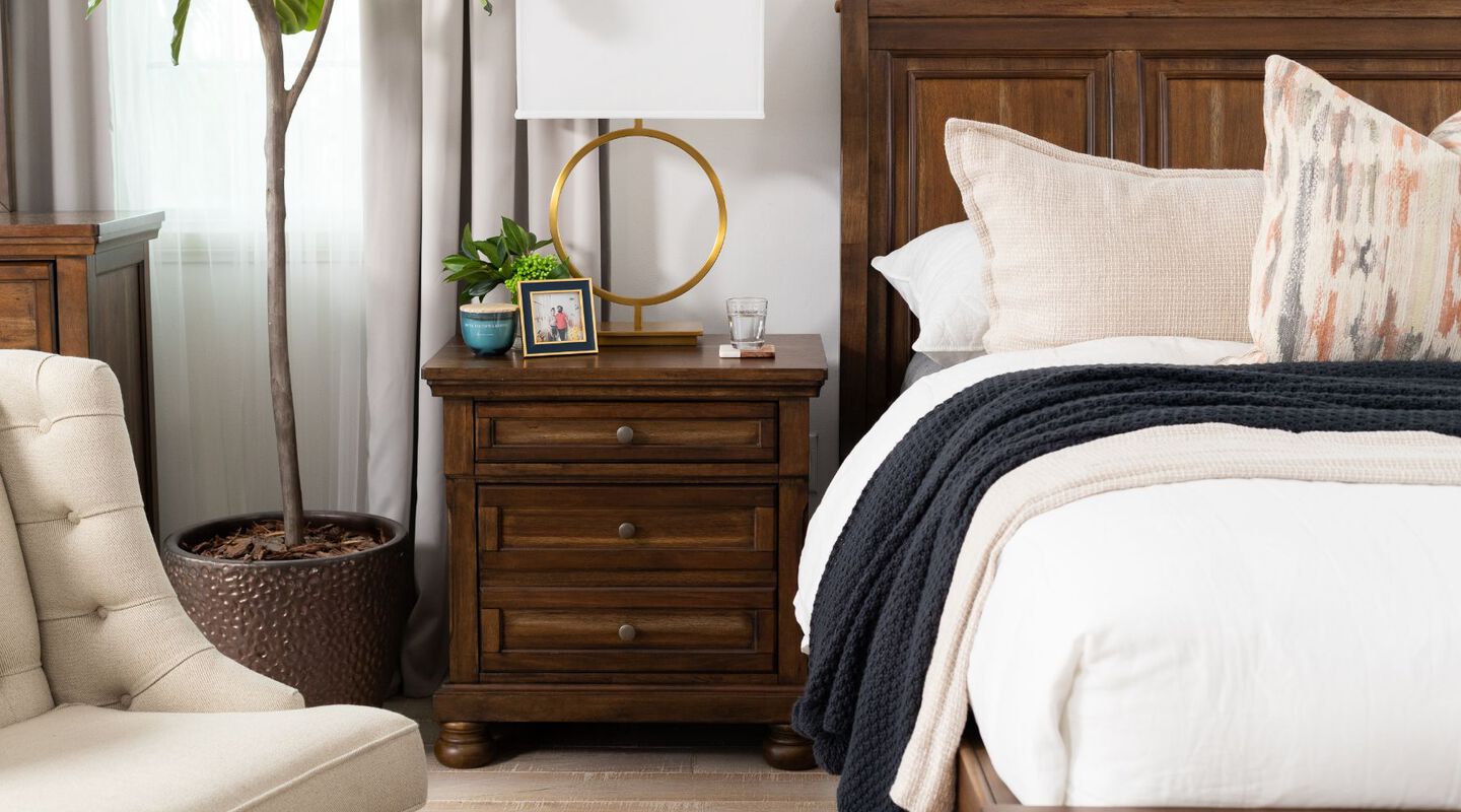 Bedroom with beige chair, wooden bed with white bedding, and a dark wooden nightstand with a gold lamp on top