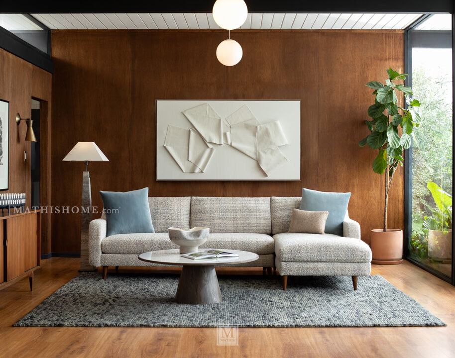 Modern sofa with a chaise, upholstered in a subtle light gray and white pattern, complemented by blue and tan pillows and supported by wooden legs.